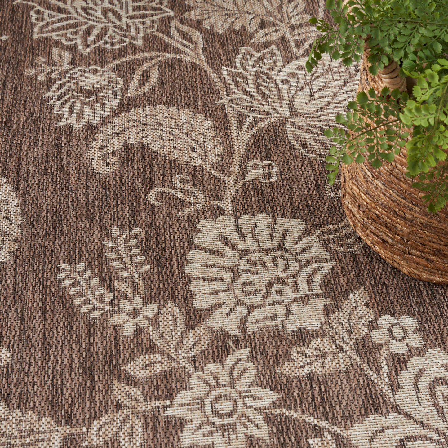 Brown woven rug with large beige floral pattern and natural texture; potted plant in a wicker basket beside.