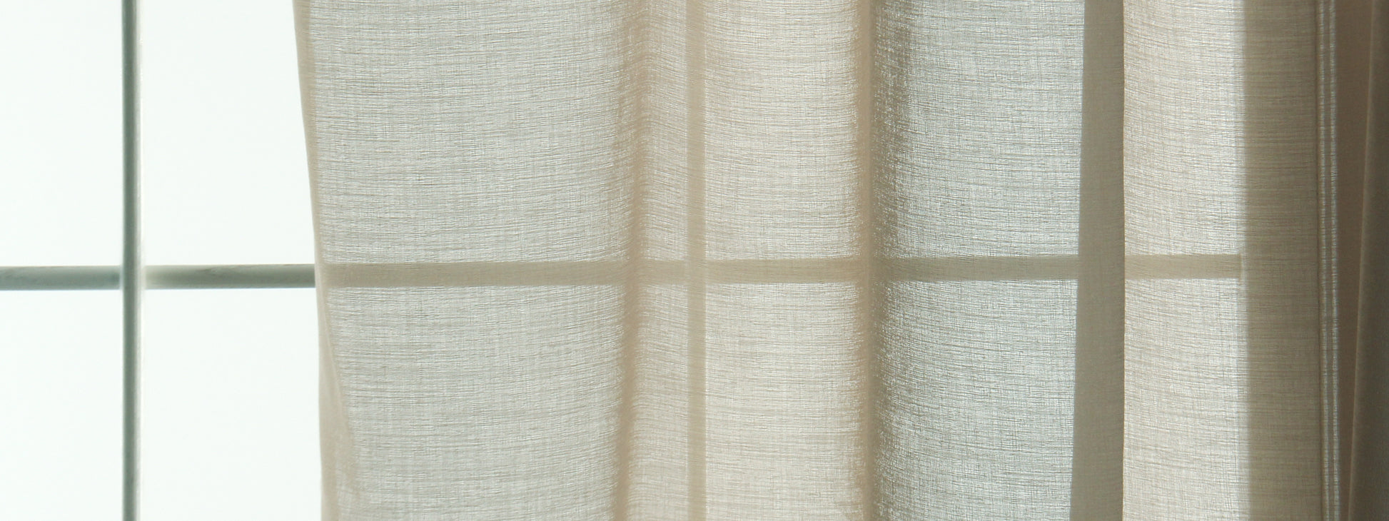 Sand-colored sheer curtains with linen texture hanging by a window, letting soft daylight through.