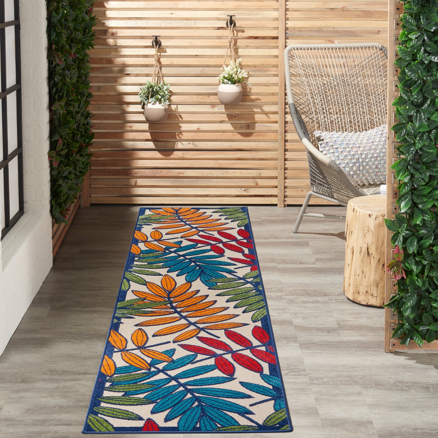 Outdoor wooden patio with a long tropical multicolor leaf-pattern rug (orange, blue, green, red), wicker chair, and hanging planters.