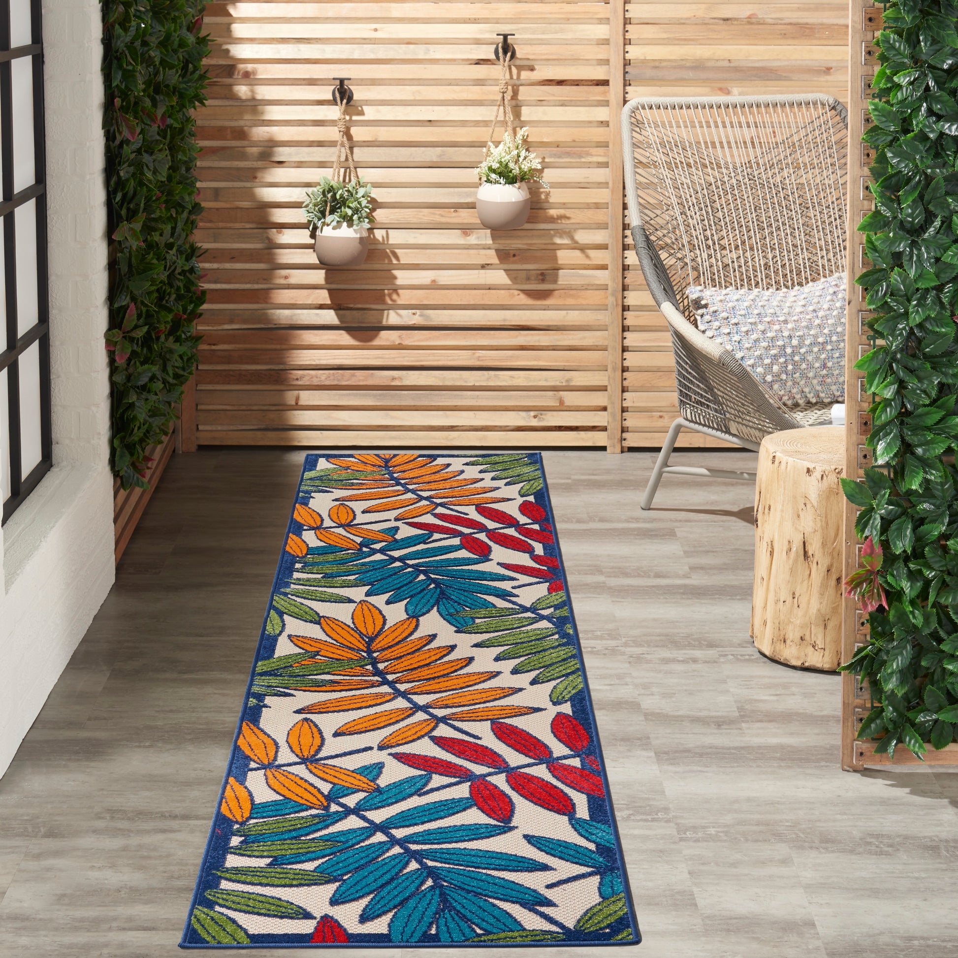 Outdoor wooden patio with a long tropical multicolor leaf-pattern rug (orange, blue, green, red), wicker chair, and hanging planters.