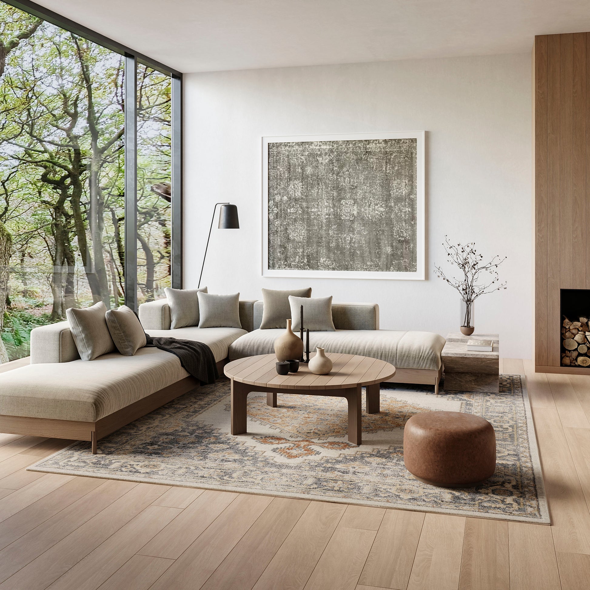 Neutral living room with beige sectional, round wood coffee table, muted cushions, patterned rug, and large windows.