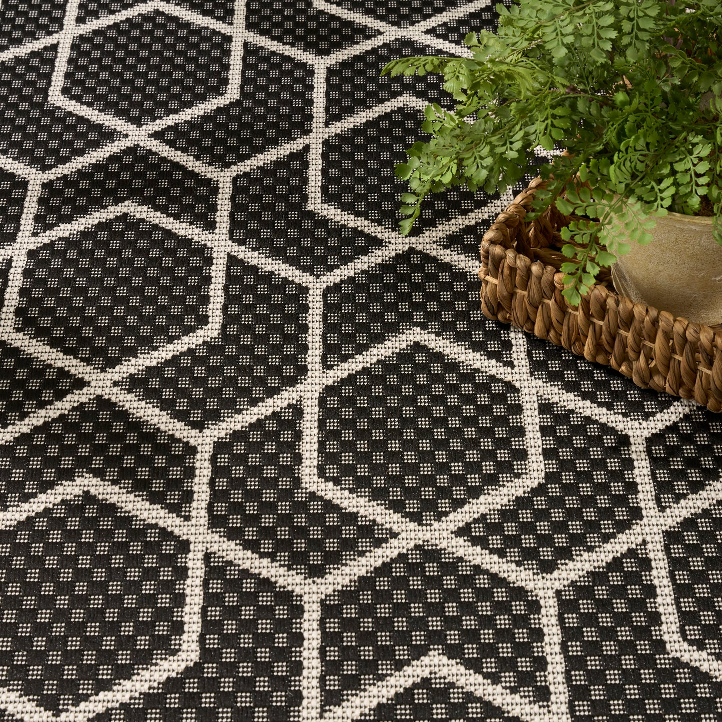 Black woven rug with white hexagon pattern and dotted interior; plant in wicker basket rests on its edge.
