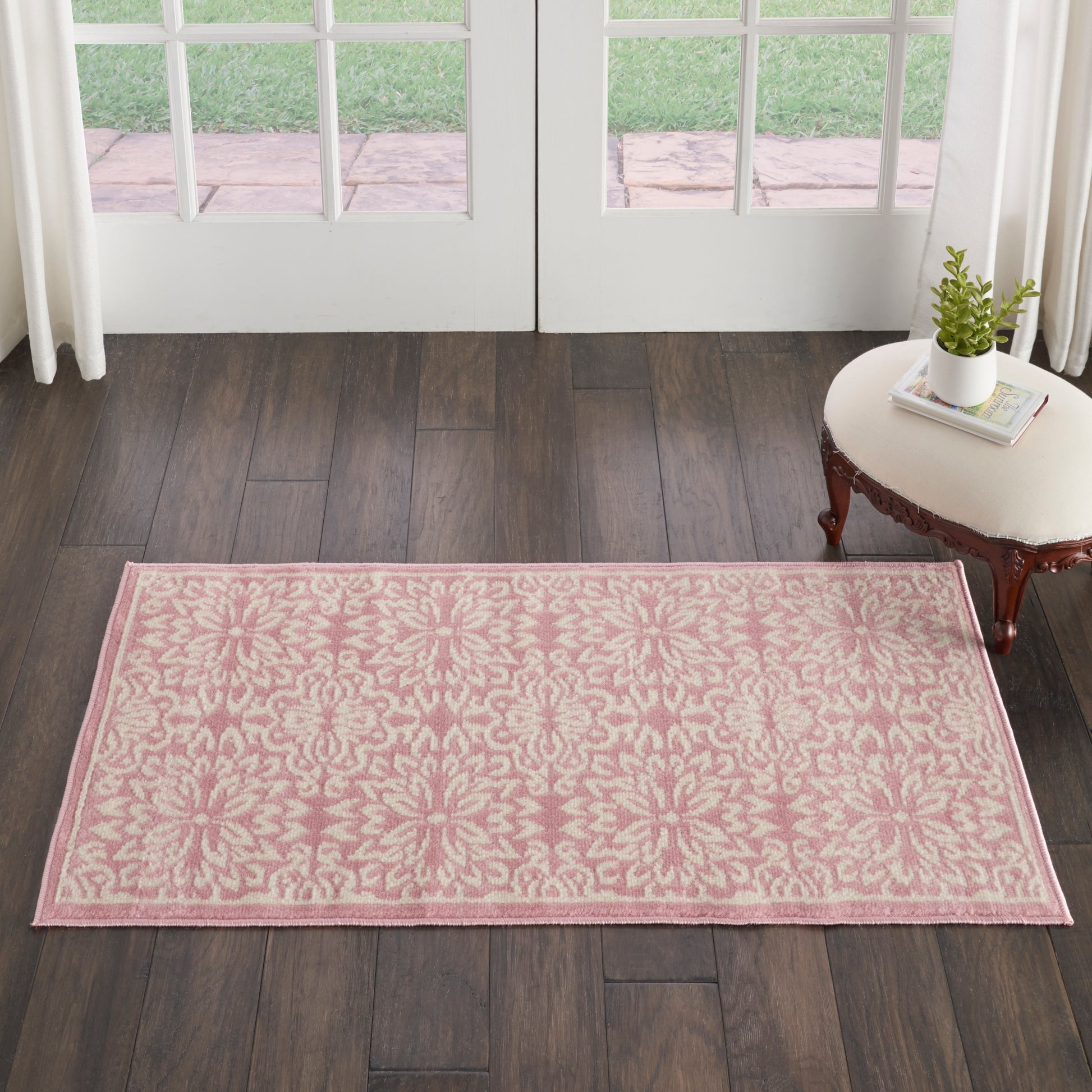 Rectangular pink rug with cream damask pattern on dark wood floor near French doors, beside a small side table with plant.