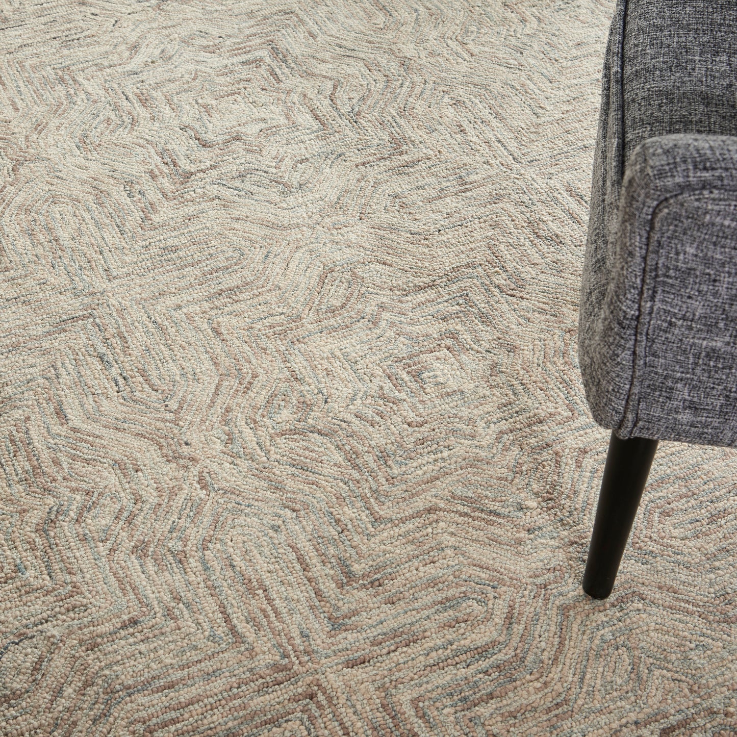 Beige textured rug with abstract, wavy pattern; partial view of a gray upholstered chair leg on the right
