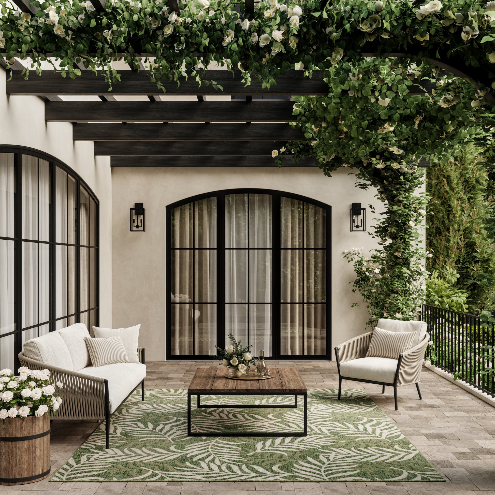 Beige stucco patio with arched doors, cream cushioned seating, wooden coffee table, palm-leaf rug, greenery overhead.