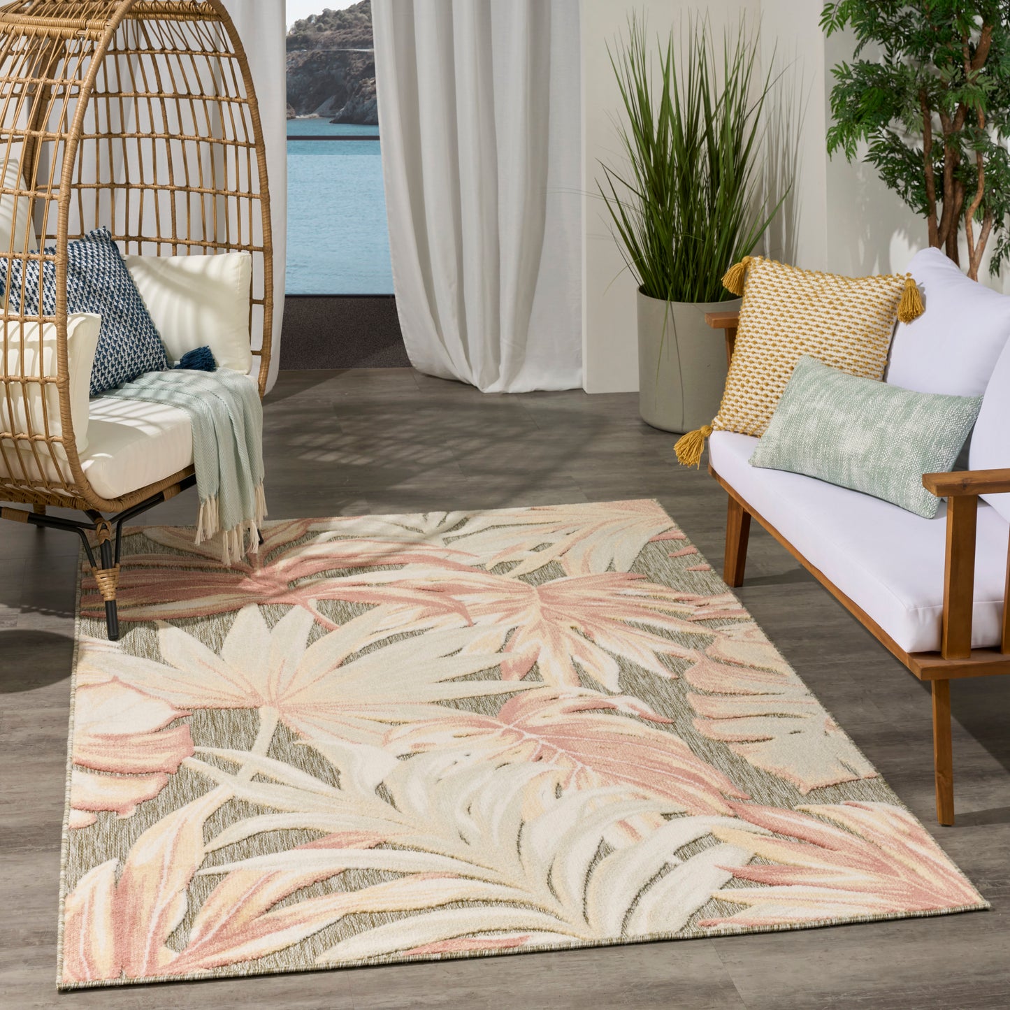 Pink-toned tropical leaf rug on dark wood floor with wicker hanging chair and white sofa in a sunlit seaside living area.