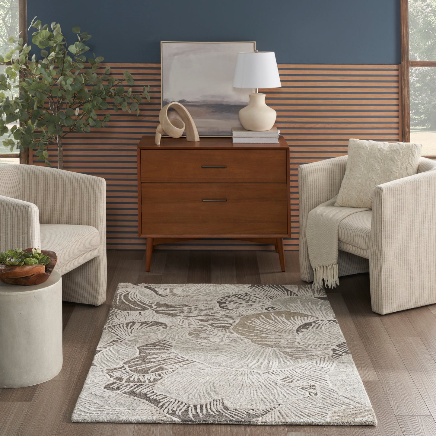 Mid-century living room: warm wood dresser, beige upholstered armchairs, palm rug, blue wall with wood slats, plant, lamp.