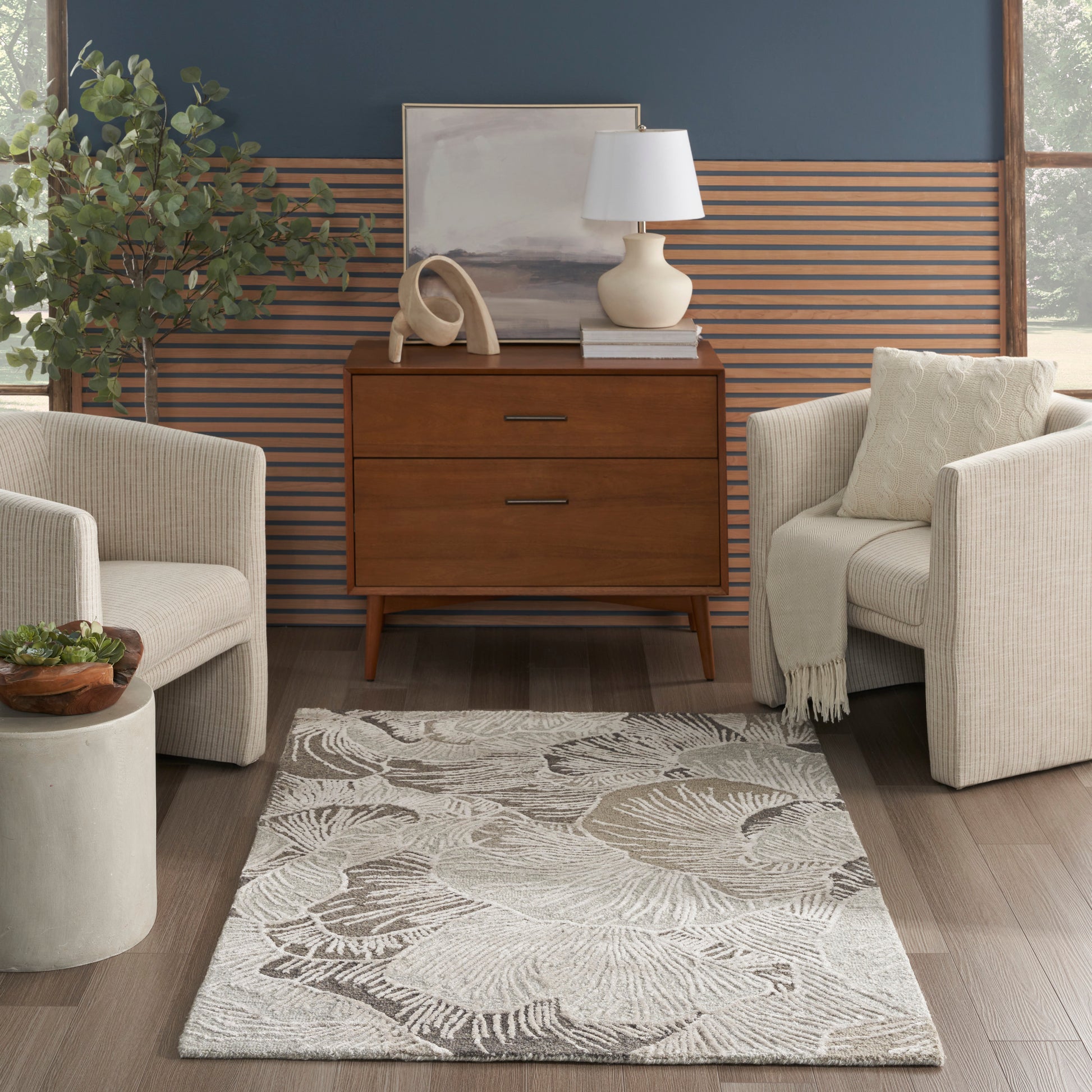 Mid-century living room: warm wood dresser, beige upholstered armchairs, palm rug, blue wall with wood slats, plant, lamp.