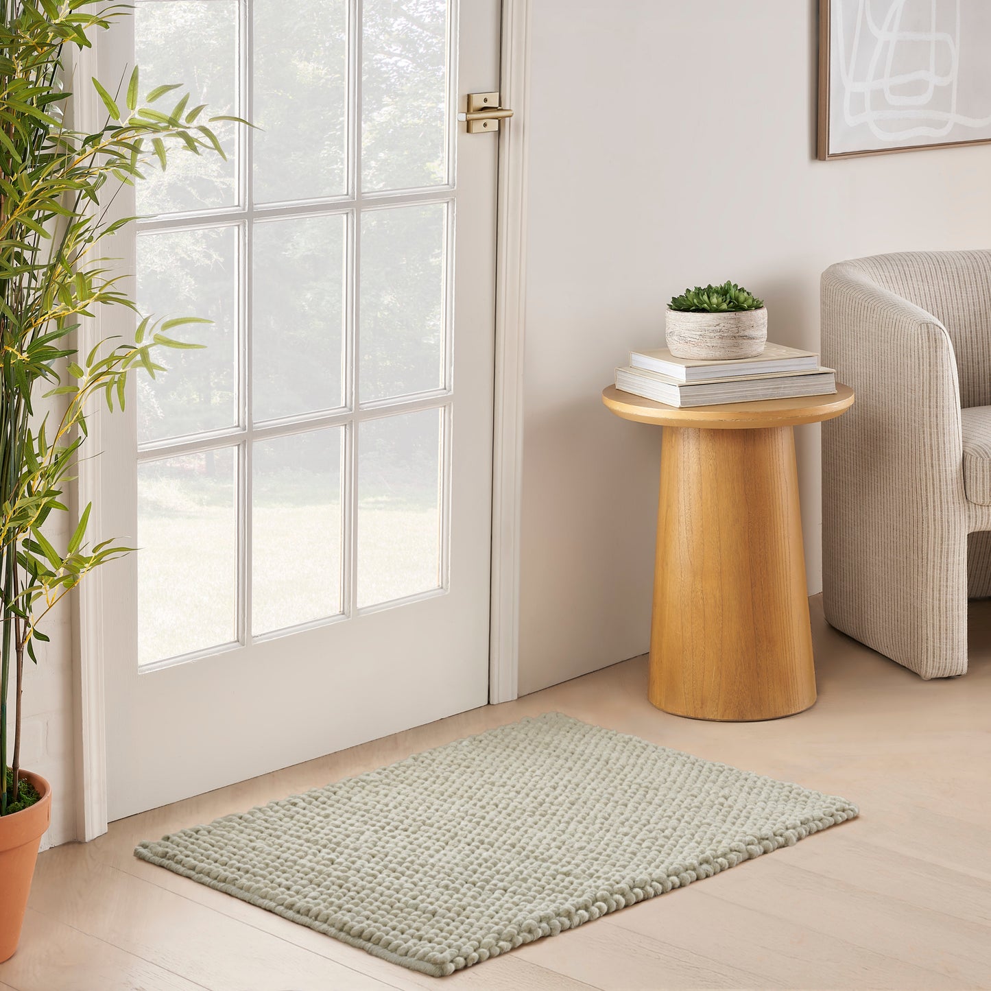 Cozy entryway: round wooden side table with books and plant, beige sofa, glass-pane door, and textured sage rug.