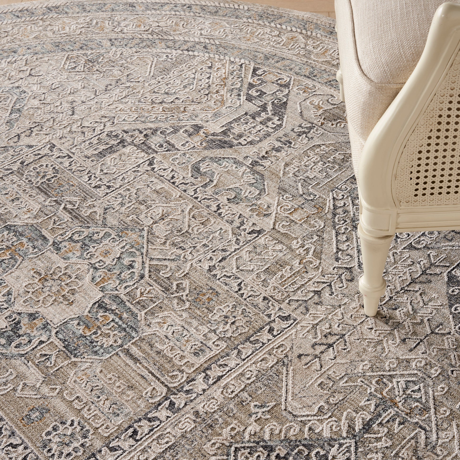 Neutral gray round oriental rug with intricate geometric and floral motifs; beige cane-chair leg resting on the rug's edge.