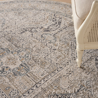 Neutral gray round oriental rug with intricate geometric and floral motifs; beige cane-chair leg resting on the rug's edge.