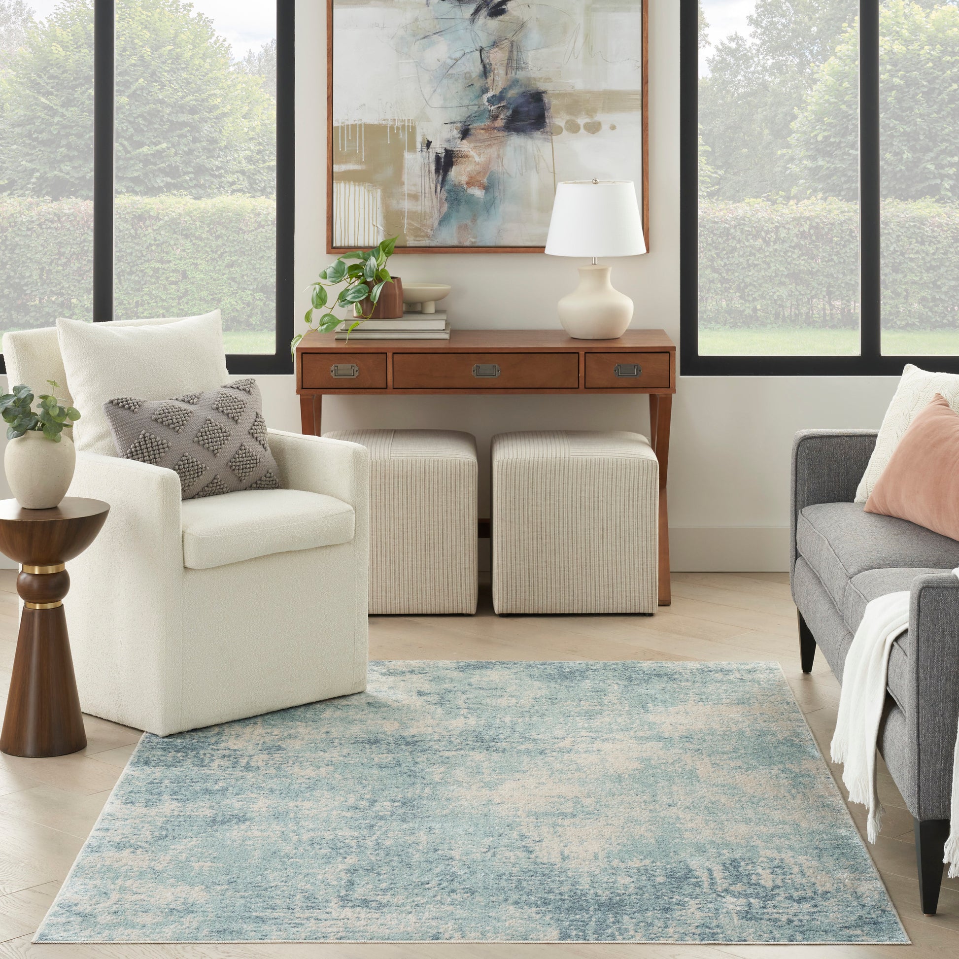 Bright living room with white armchair, grey sofa, blue distressed rug, wooden console with drawers, beige ottomans, art.