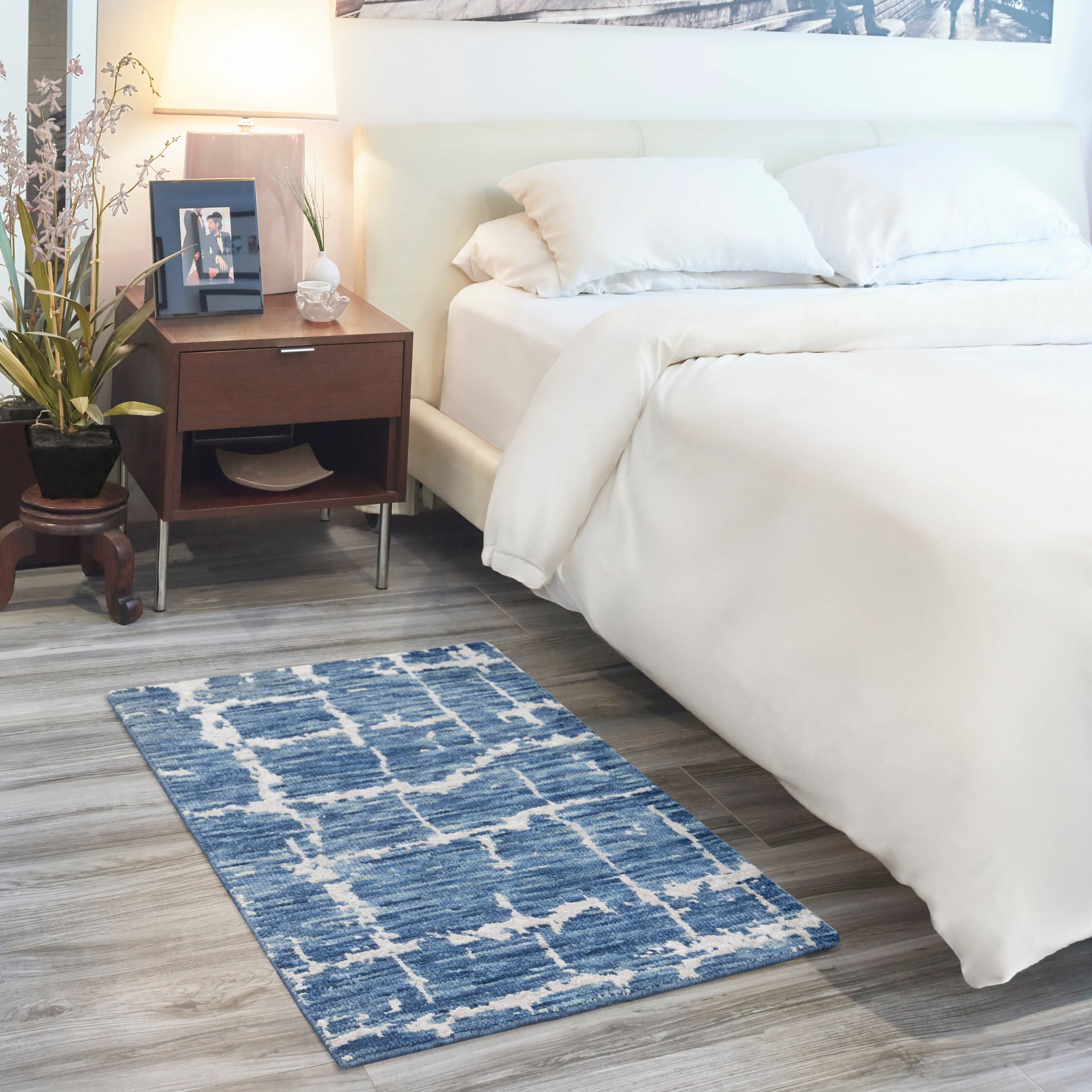 Blue geometric rug on light wood floor beside a white bed; dark wood nightstand with lamp, plant and photo frame.