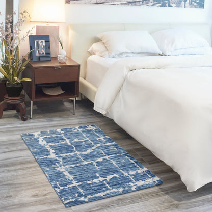 Blue geometric rug on light wood floor beside a white bed; dark wood nightstand with lamp, plant and photo frame.