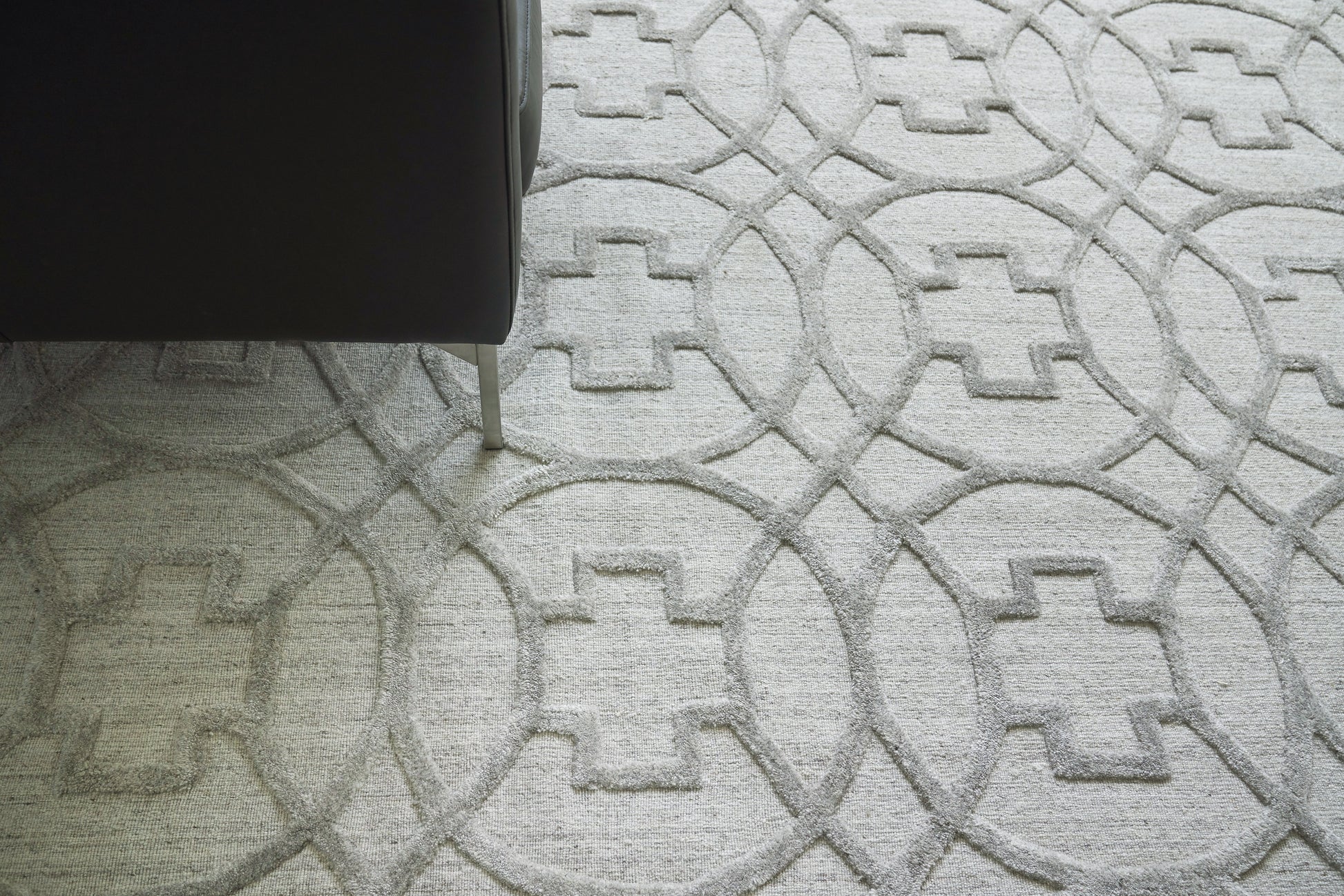 Partial view of a light gray geometric rug with interlocking circle and square patterns, dark sofa leg on the left.