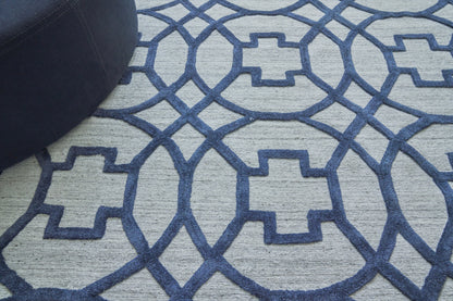 Blue geometric trellis rug on light gray field; flat-weave, modern pattern; dark blue round ottoman visible in corner.