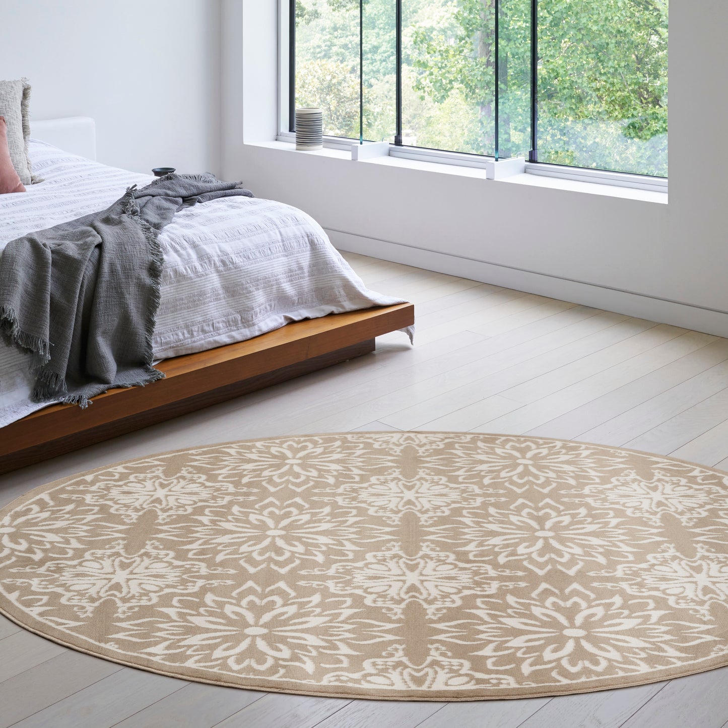 Round beige rug with white floral pattern in a modern bedroom beside a low wooden bed with white linens and gray throw.