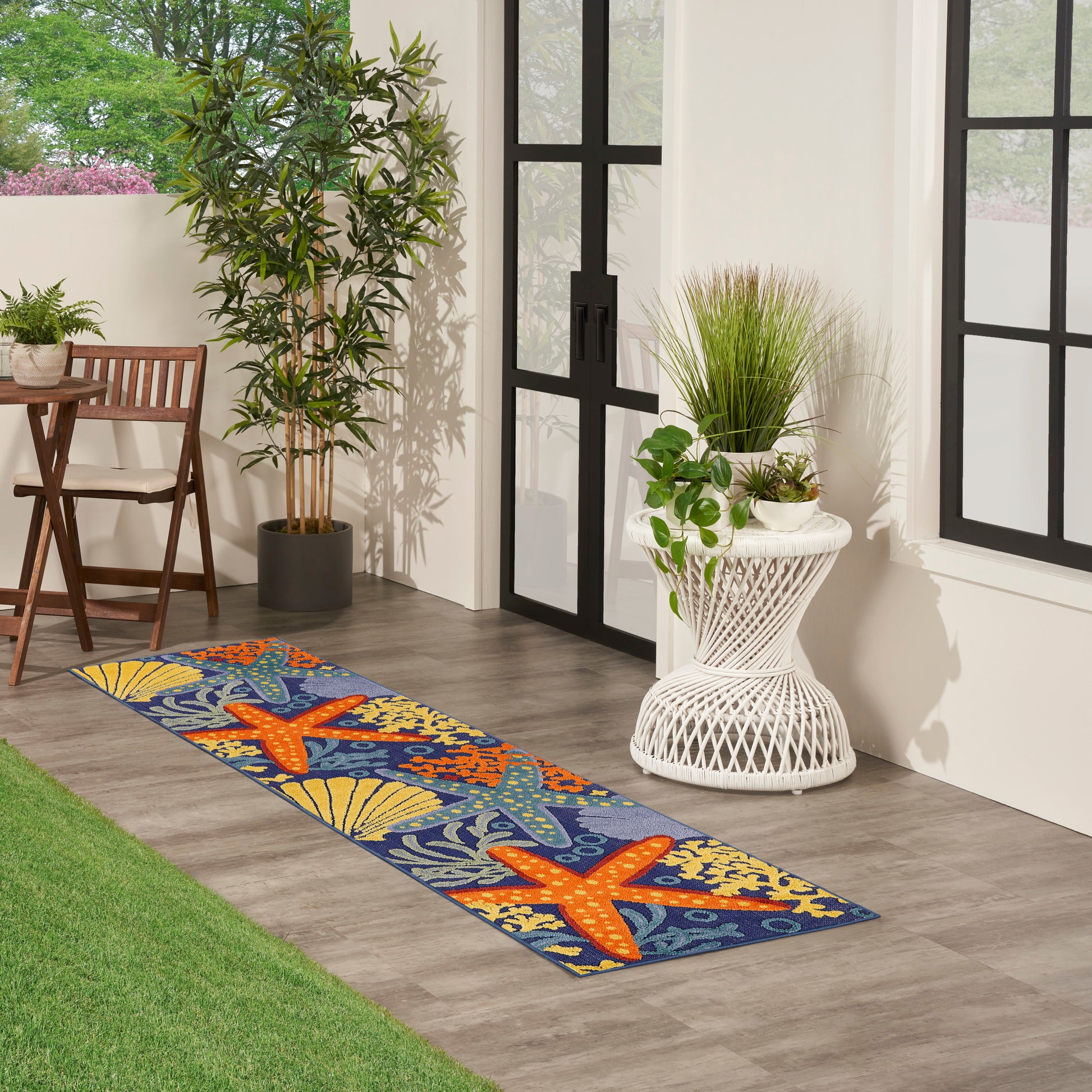 Coastal outdoor rug runner with orange starfish and seashell motifs on a blue background, laid on a sunlit patio with potted plants.
