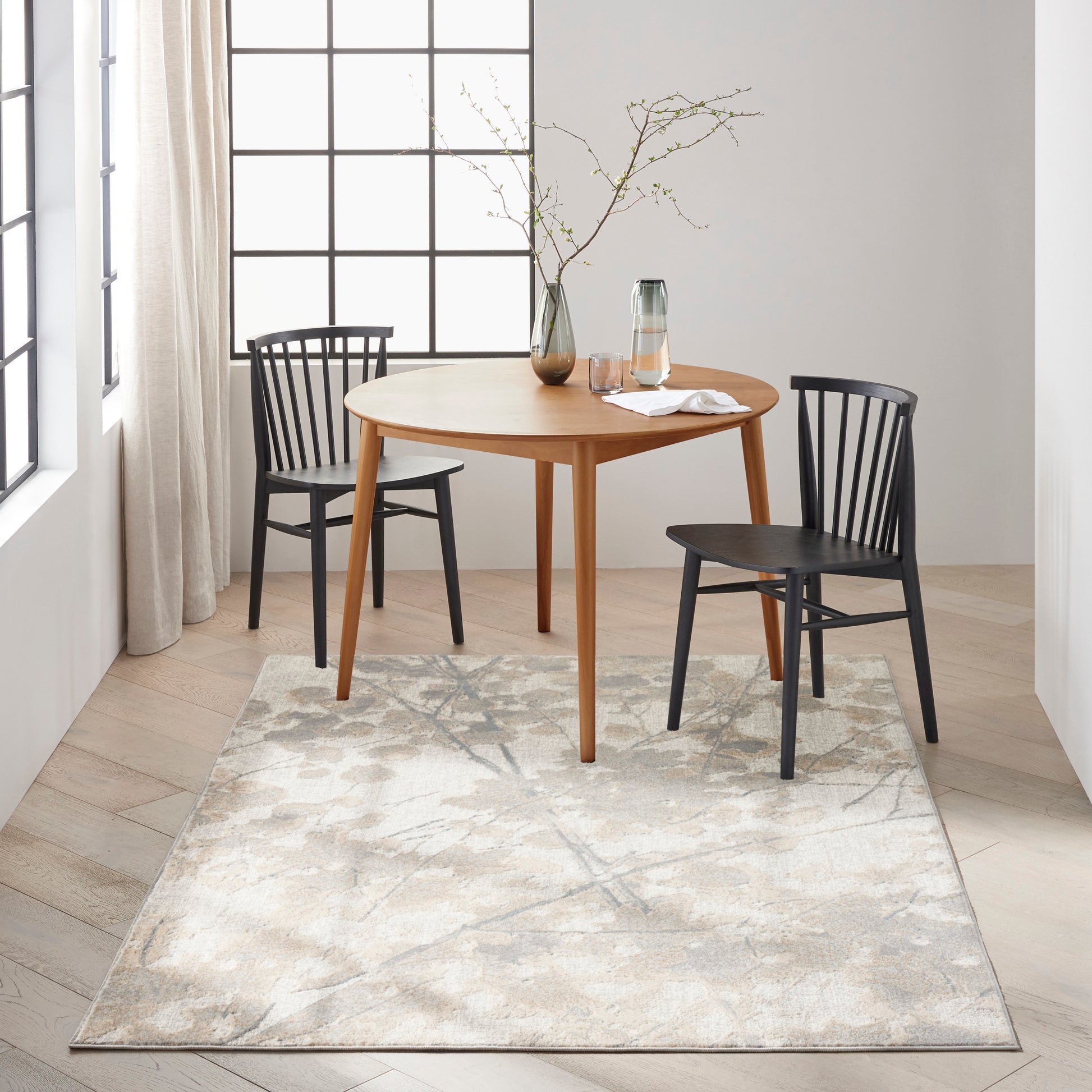 Light patterned rug beneath a round wooden dining table with two black chairs, a glass carafe, and a vase with branches by grid windows.
