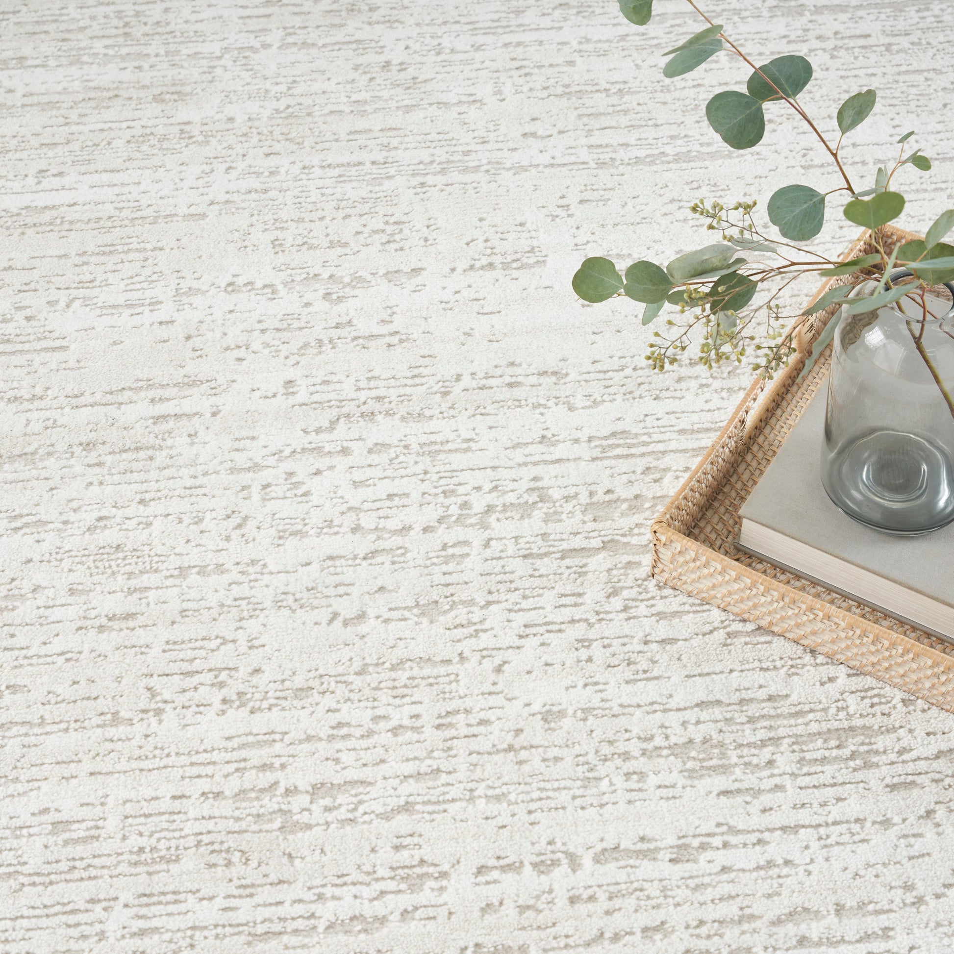 Beige textured rug backdrop with a woven wicker tray holding a glass vase, books & eucalyptus sprigs.