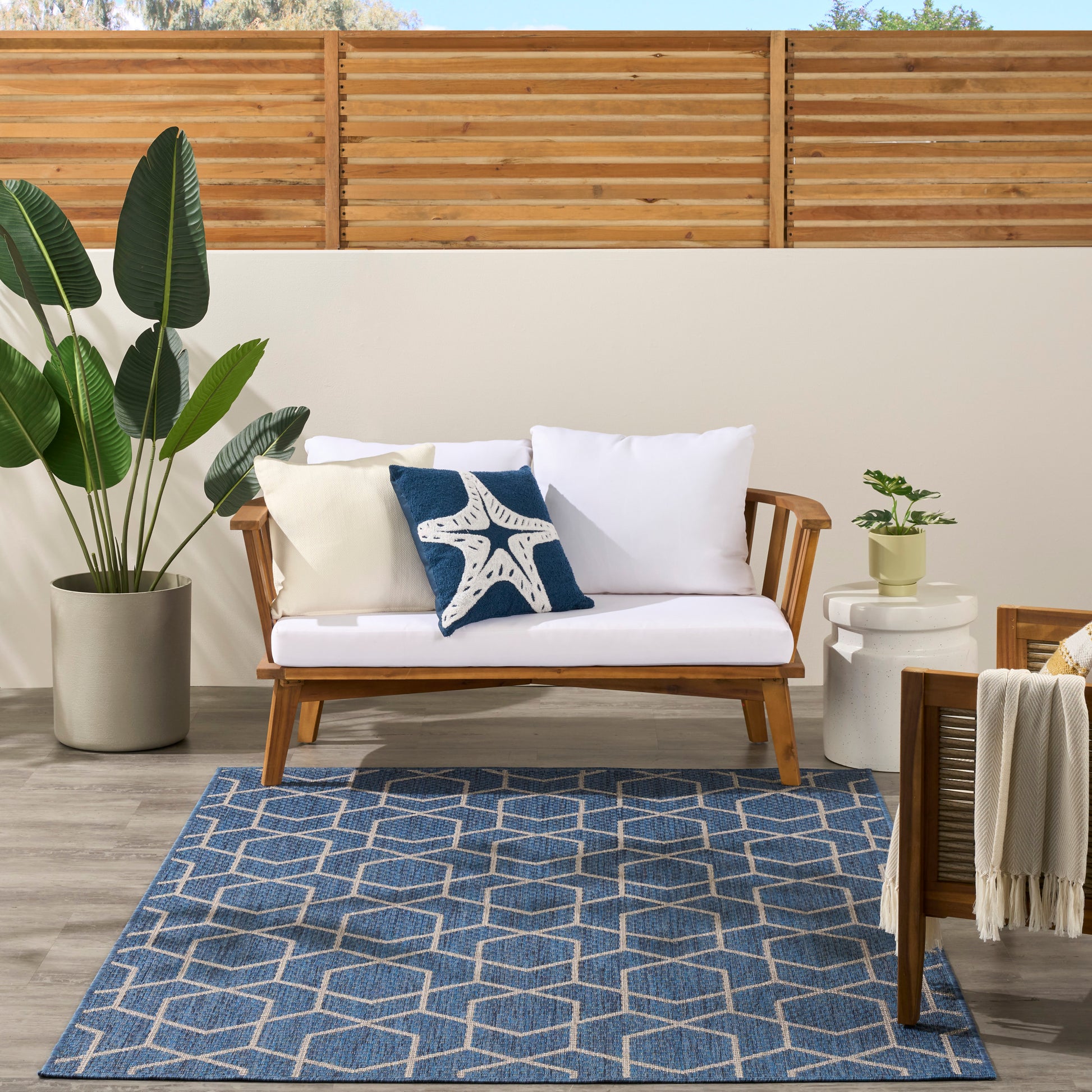 Outdoor wooden sofa with white cushions and blue starfish pillow, potted greenery, geometric blue rug, wooden fence backdrop.