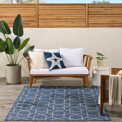 Outdoor wooden sofa with white cushions and blue starfish pillow, potted greenery, geometric blue rug, wooden fence backdrop.