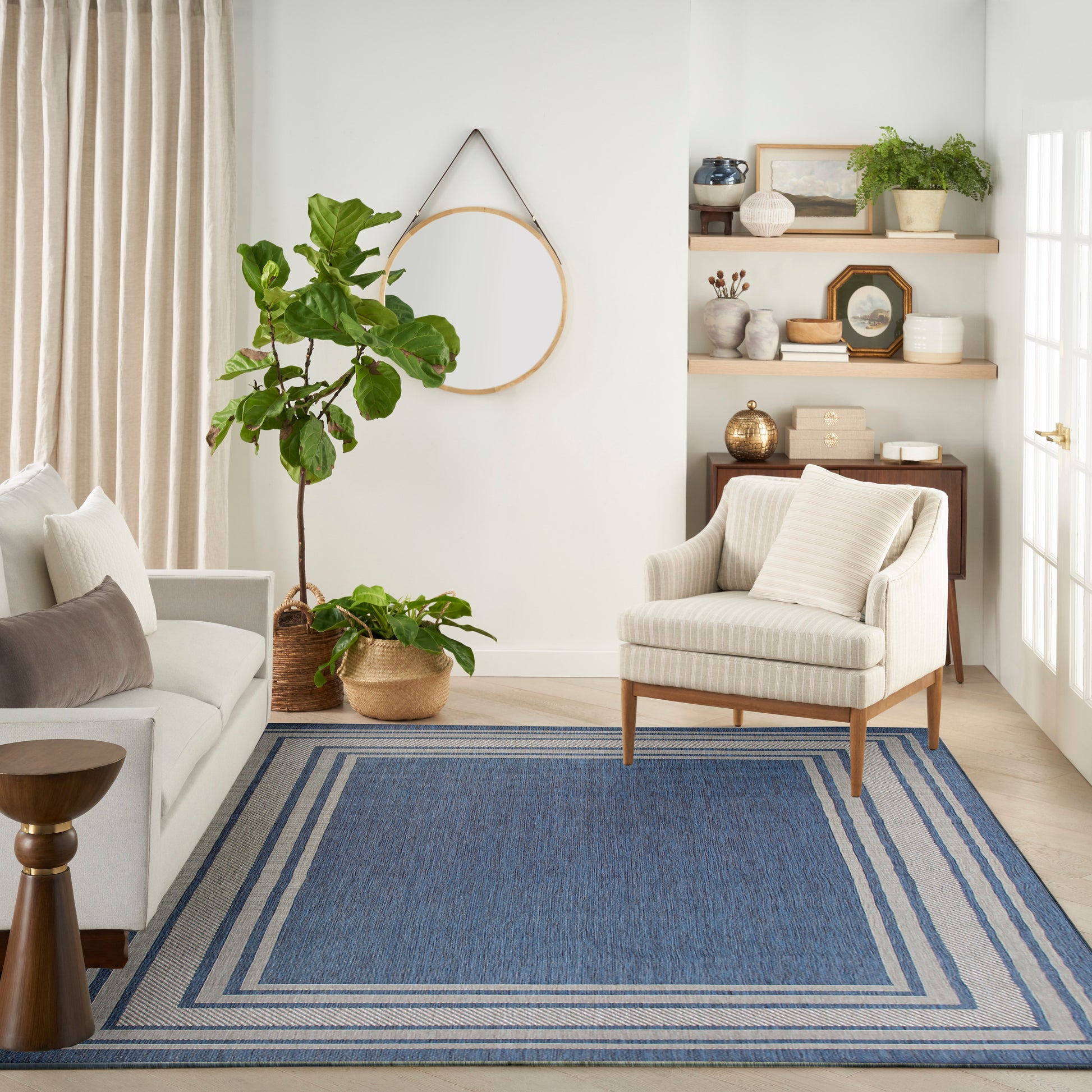 Cozy living room with beige sofa, striped armchair, blue bordered area rug, and wood shelves with plants.