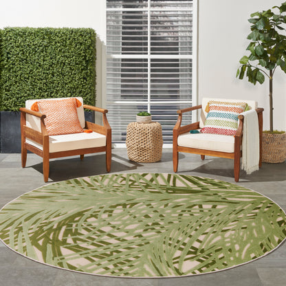 Outdoor patio: round green palm-leaf rug, wood chairs with cream cushions, colorful pillows, woven side table, plants.