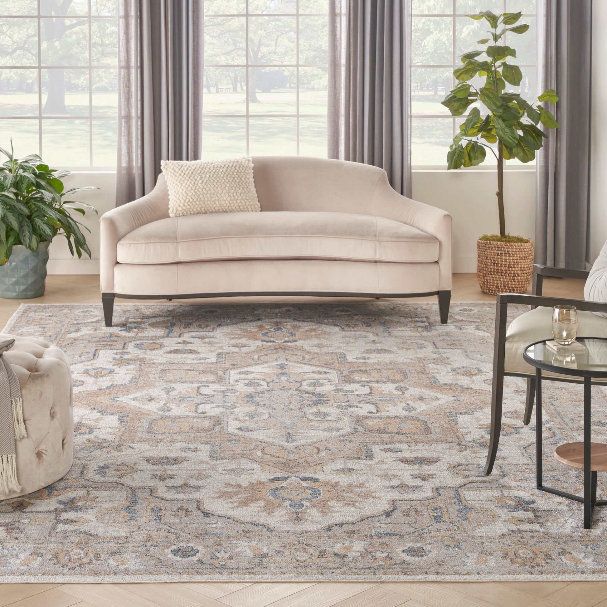 Soft beige curved sofa with dark legs, patterned rug, green plants, gray curtains, and glass coffee table.