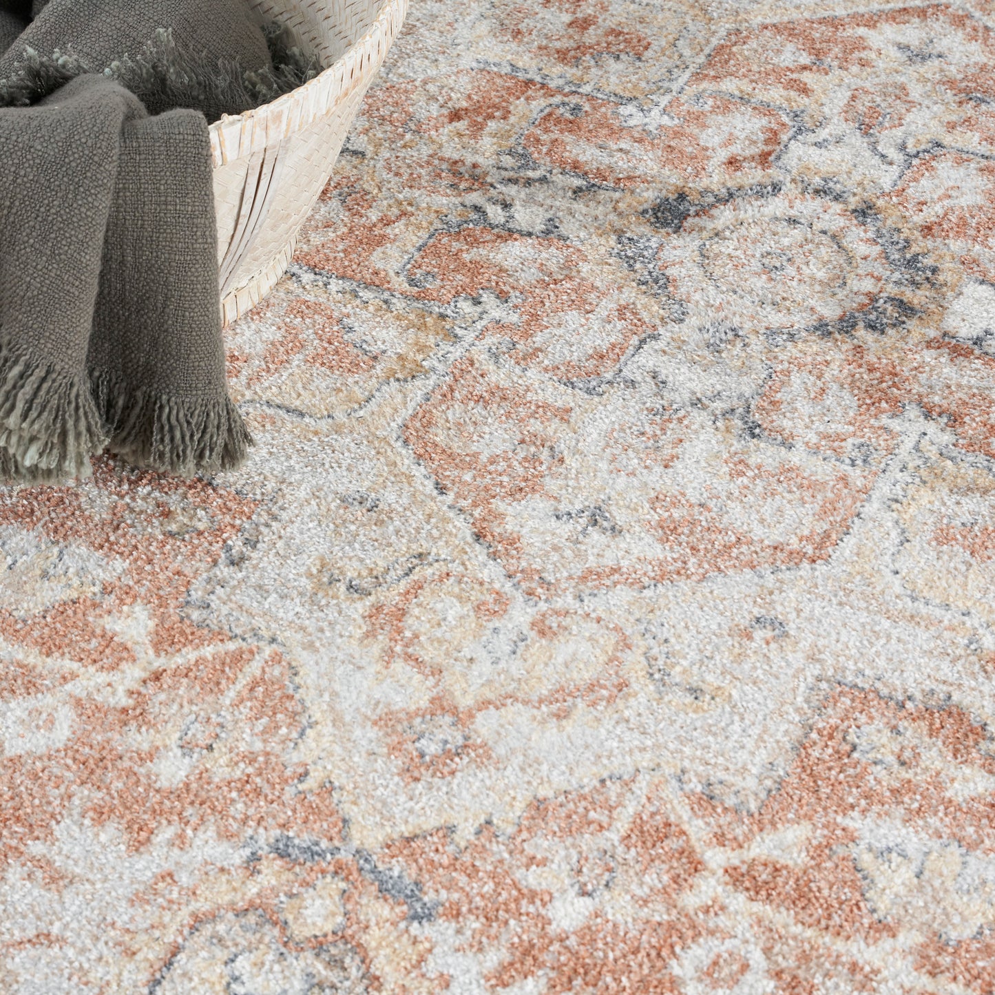 Patterned area rug in orange, cream and gray with fringe; wicker basket with a dark gray throw.