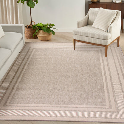 Beige textured area rug with double border, cream armchair on wooden legs, and potted plants in woven baskets in a neutral living room.