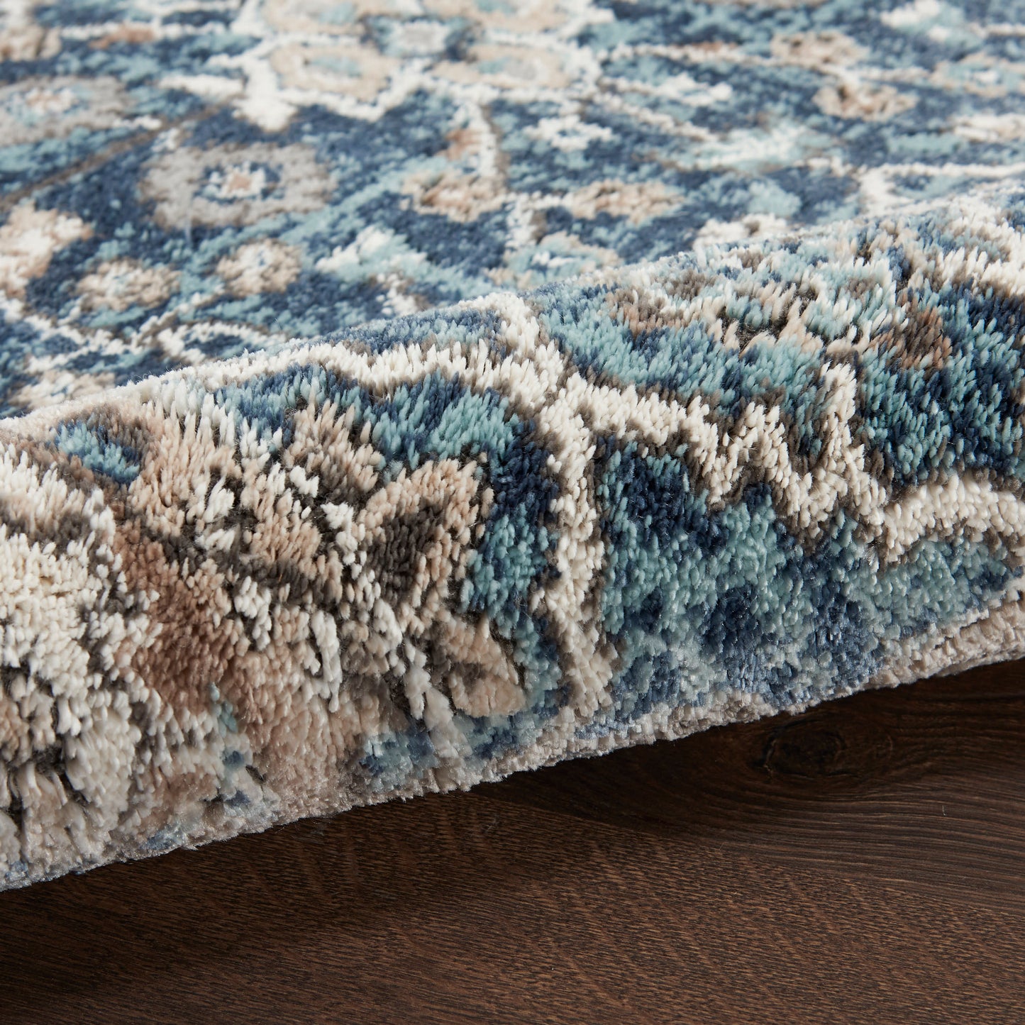 Close-up view of a plush blue, teal, beige patterned rug with a shaggy edge on dark wood floor.