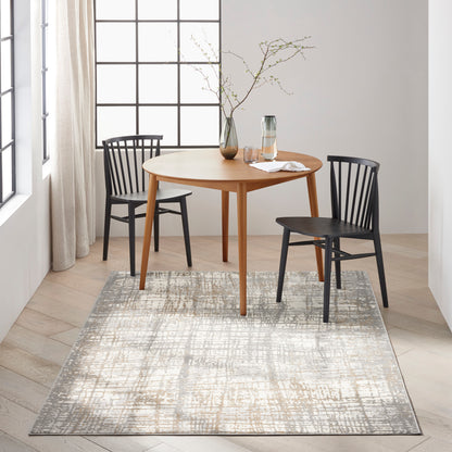 8' x 10' modern rug with a gray abstract pattern; a round light-wood dining table and two black Windsor chairs sit on it, with a vase of branches and a glass carafe nearby.