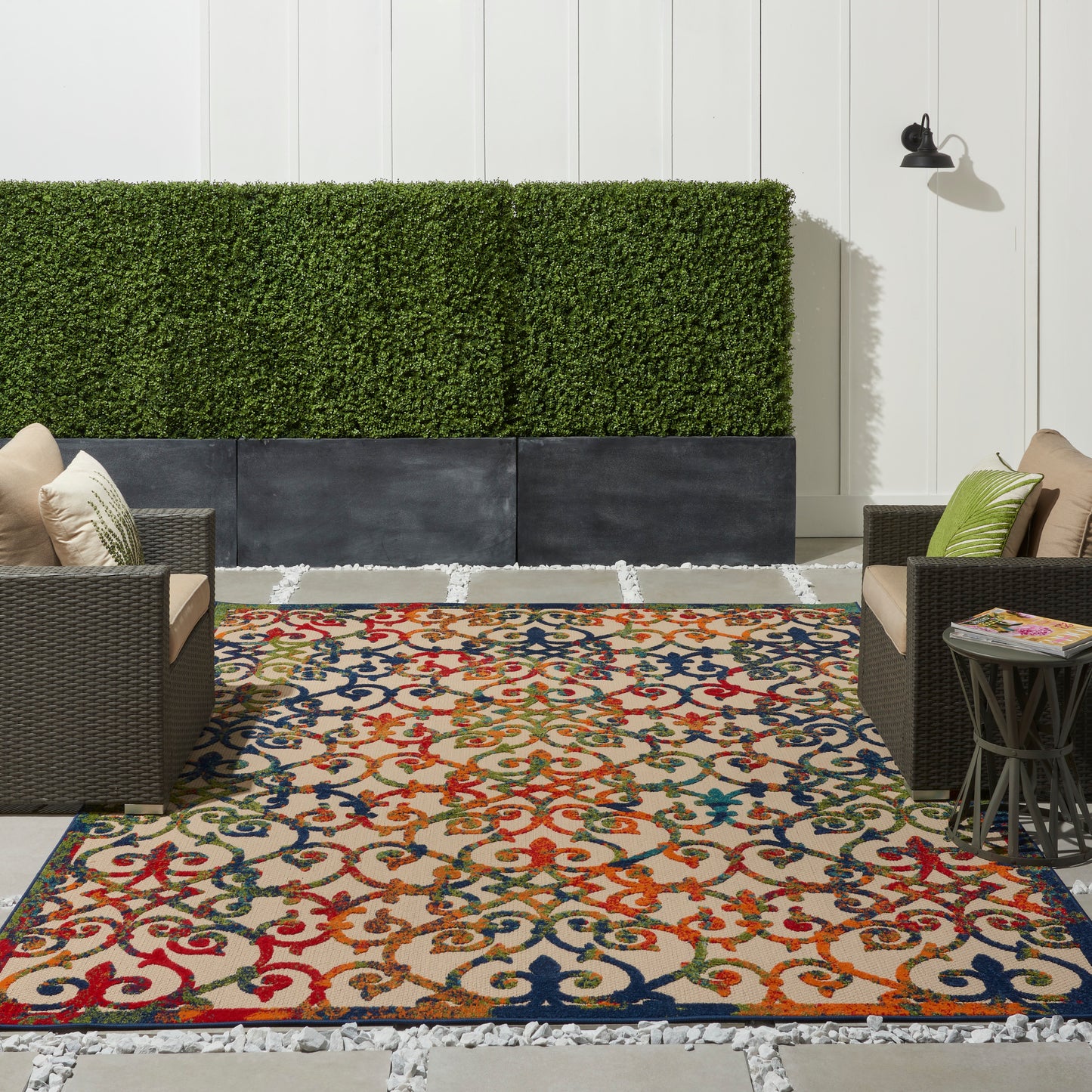 Vibrant outdoor rug with red, blue, orange and green scroll pattern, flanked by brown wicker chairs and a greenery hedge.