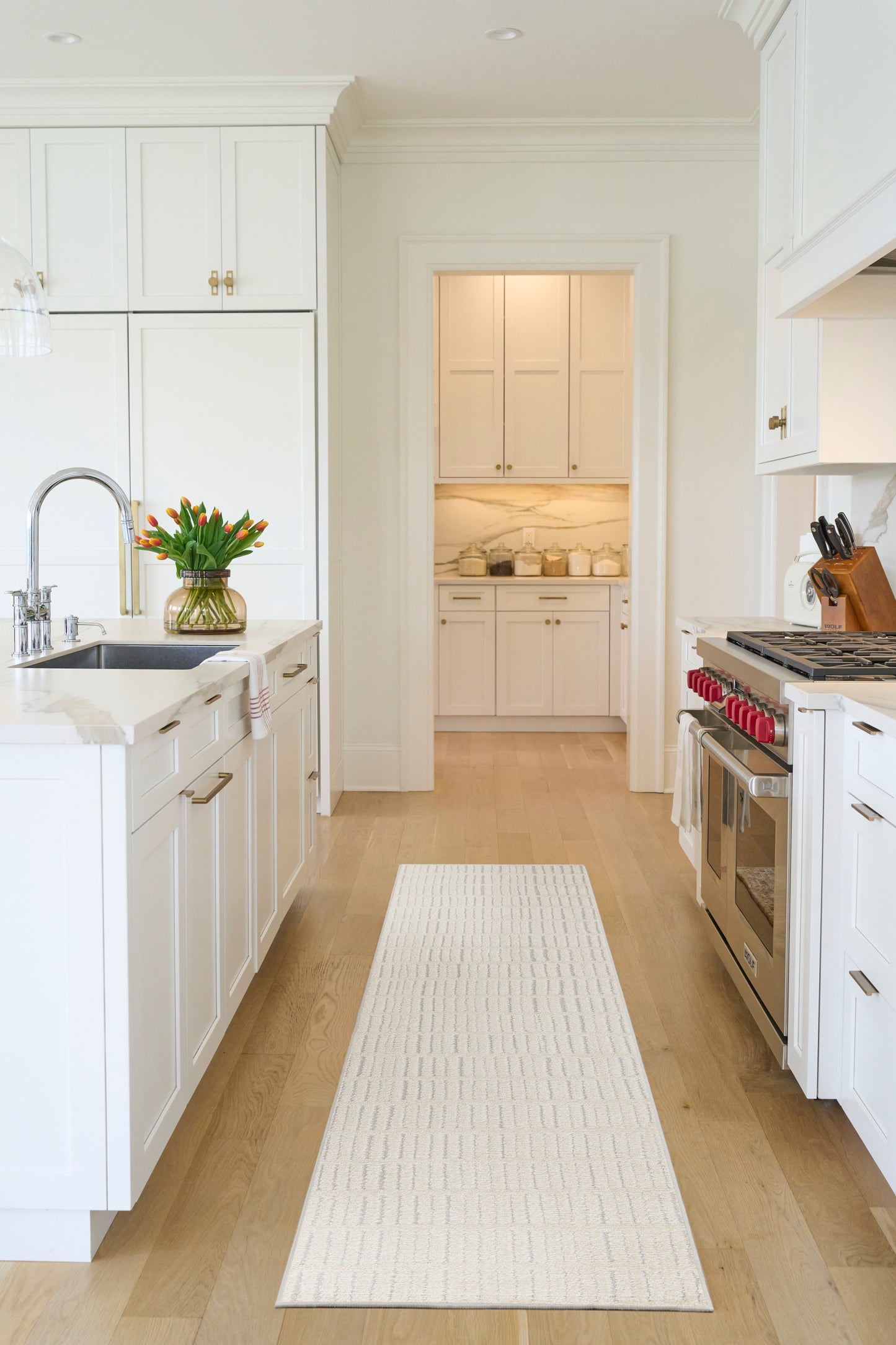 Bright white kitchen with marble countertops, stainless gas range, long white runner rug, and tulips on the island.