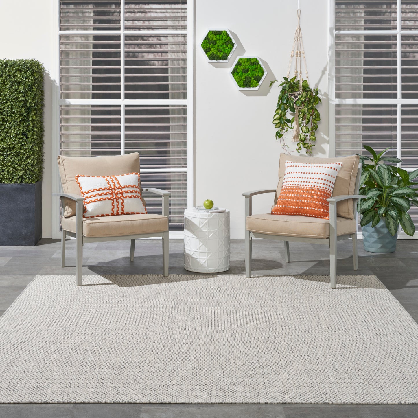Two beige patio chairs with orange pillows flank a white side table with a green apple, on a light rug amid greenery.