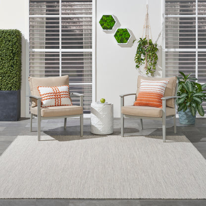 Two beige patio chairs with orange pillows flank a white side table with a green apple, on a light rug amid greenery.