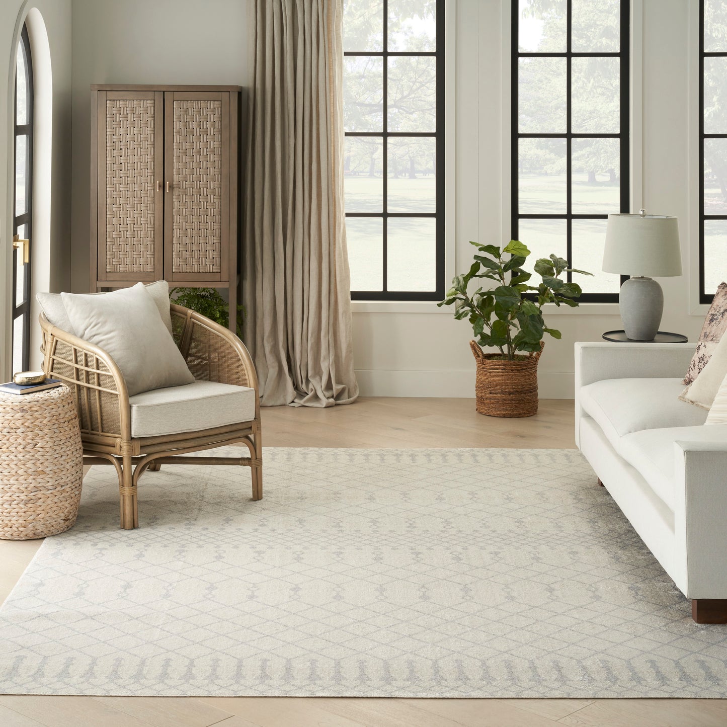 Neutral rug with a pale geometric pattern in a living room setting, shown with a white sofa, rattan armchair, woven cabinet, large windows, and a potted plant.