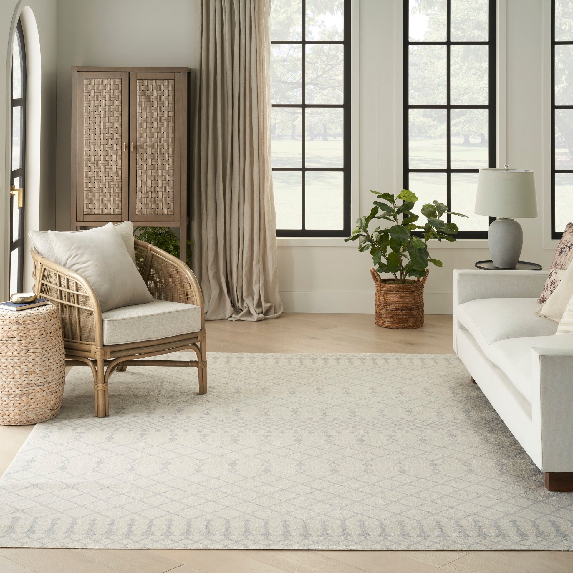Neutral rug with a pale geometric pattern in a living room setting, shown with a white sofa, rattan armchair, woven cabinet, large windows, and a potted plant.