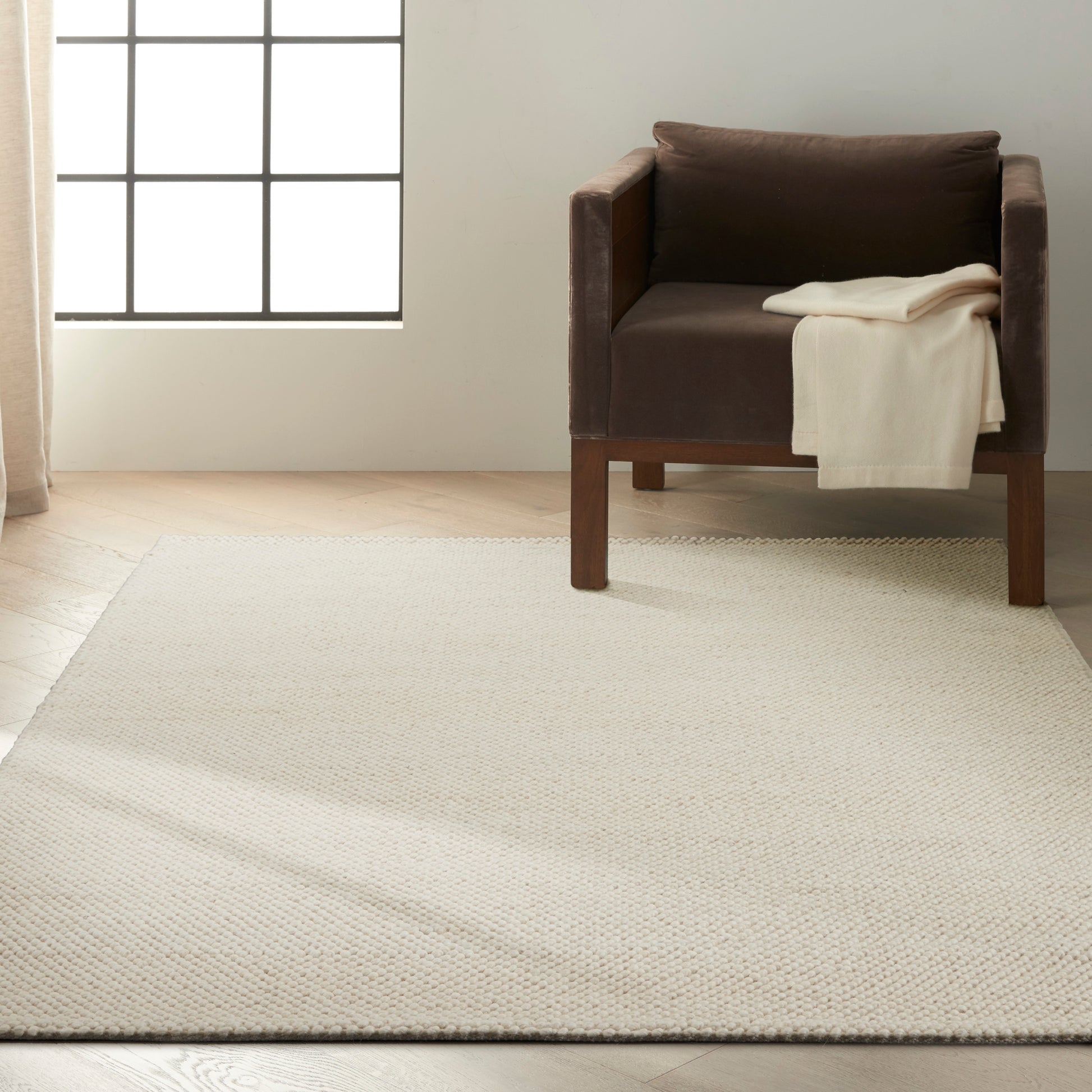 Brown leather armchair with wooden legs, cream blanket draped, on a large beige woven area rug by a grid window.