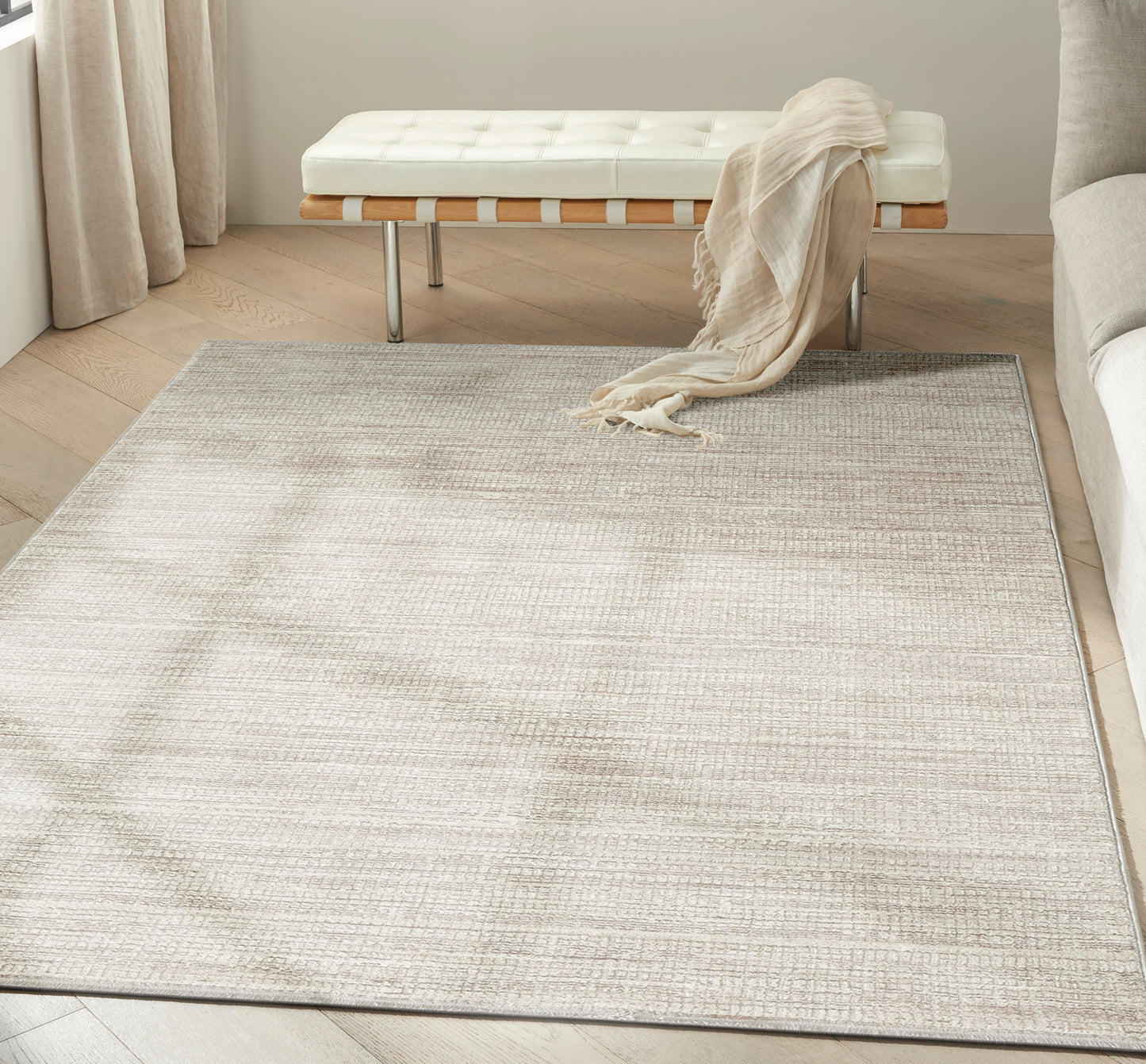 Neutral beige woven area rug in a light living room with a white tufted bench, beige throw, and light wood floors.