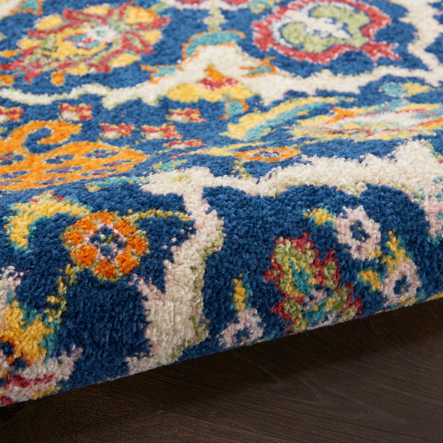 Vibrant blue area rug close-up with multicolor floral motifs, plush pile and cream border on dark wood floor.