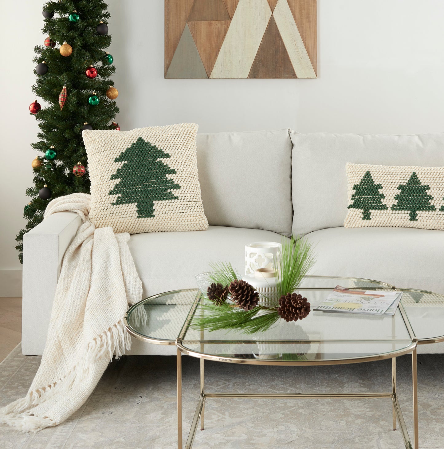 Cozy living room with Christmas tree, beige sofa, knitted green-tree cushions, throw, glass coffee table with pinecones.