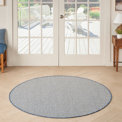 Round blue-and-white woven rug (5' diameter) on light wood floor by French doors; living room with a blue chair, side table and lamp.