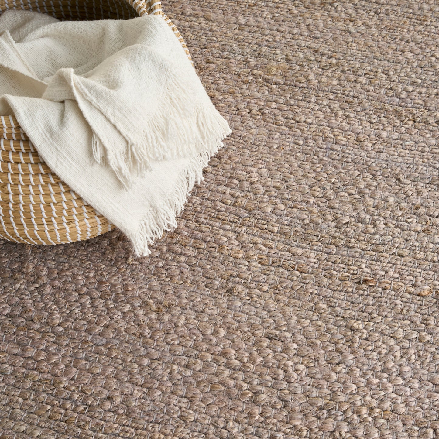 Natural tan jute rug with chunky woven texture; cream fringed blanket draped over a woven basket.