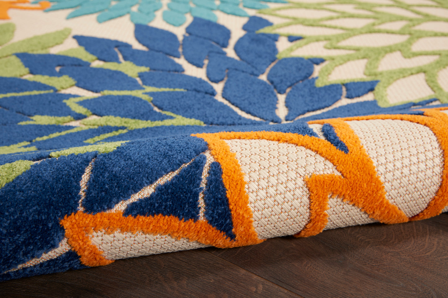 Close-up of a vibrant rug with blue, teal, lime-green floral patterns and orange edging, rolled on a dark wood floor.