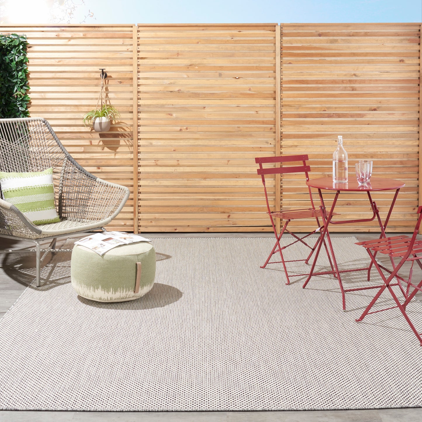 9' x 12' outdoor rug in grey tones on a patio, visible with beige wicker chair and green striped cushion, round pouf, red metal bistro set, and wooden slat fence.