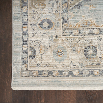 Blue, beige and ivory ornate area rug with floral medallion border and fringed edge on dark wood floor.
