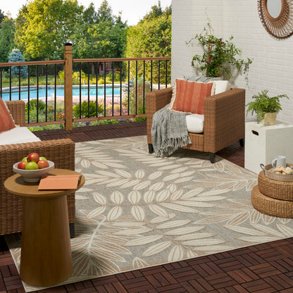 Wicker outdoor seating on leaf-pattern rug, white cushions with orange pillow, gray throw; plants, railing, pool view.