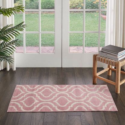 Pink rectangular area rug with white wave lattice pattern on dark hardwood floor; plant left, books on wooden bench right.